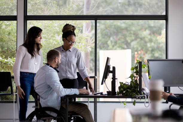 Two women and a man in a wheelchair look at a computer together in an office setting.